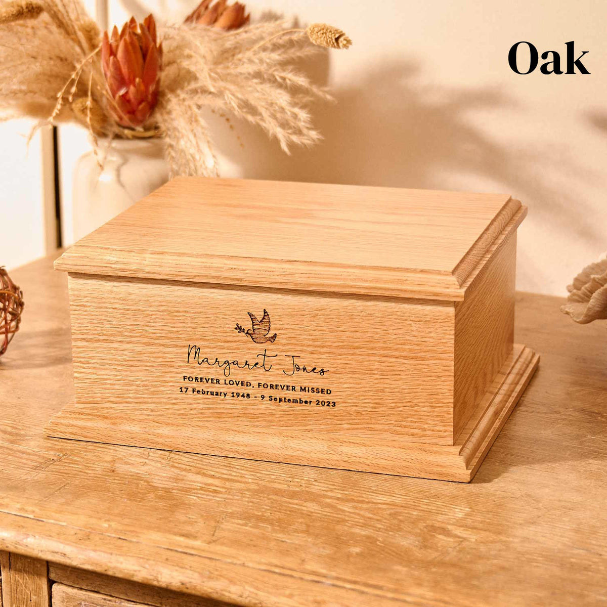 A wooden cremation urn with a personalised engraving sitting on a wooden table. The urn is made of oak. It is rectangular and has detailed bevelling on the top face and around the bottom. The engraving shows the outline of a dove in flight, carrying an olive branch in its beak. Underneath the dove a message reads "Margaret Jones - Forever Loved, Forever Missed - 17 February 1948 - 9 September 2023"