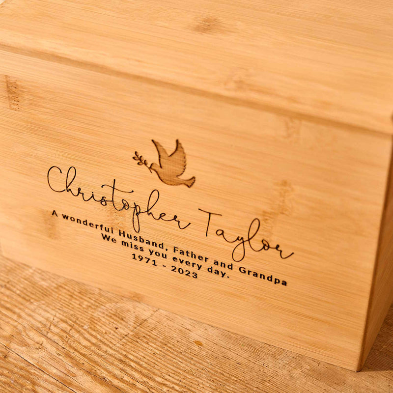 A close up photo of an engraving on a wooden cremation urn. The urn is made of light wood. The engraving shows the outline of a dove in flight, carrying an olive branch in its beak. Underneath the dove a message reads "Christopher Taylor - A wonderful husband, father and grandpa. We miss you every day - 1971-2023"