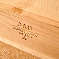 A close up of a wooden cremation urn for ashes. The urn is engraved with the words "Dad - Always Loved 1963 - 2023" and a small picture of a person sitting on a motorbike.