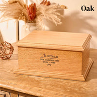 A wooden cremation urn for ashes. The cremation urn is rectangular and is made of oak. It is engraved with the words "Thomas - Our darling son - 2004-2023" and a small picture of a games console controller 