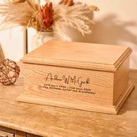 A wooden cremation urn. The urn is made light wood and has a personalised engraving which reads "Andrew W. McGrath - 14th April 1961 - 21st June 2023 - Loving Husband, Father and Grandfather