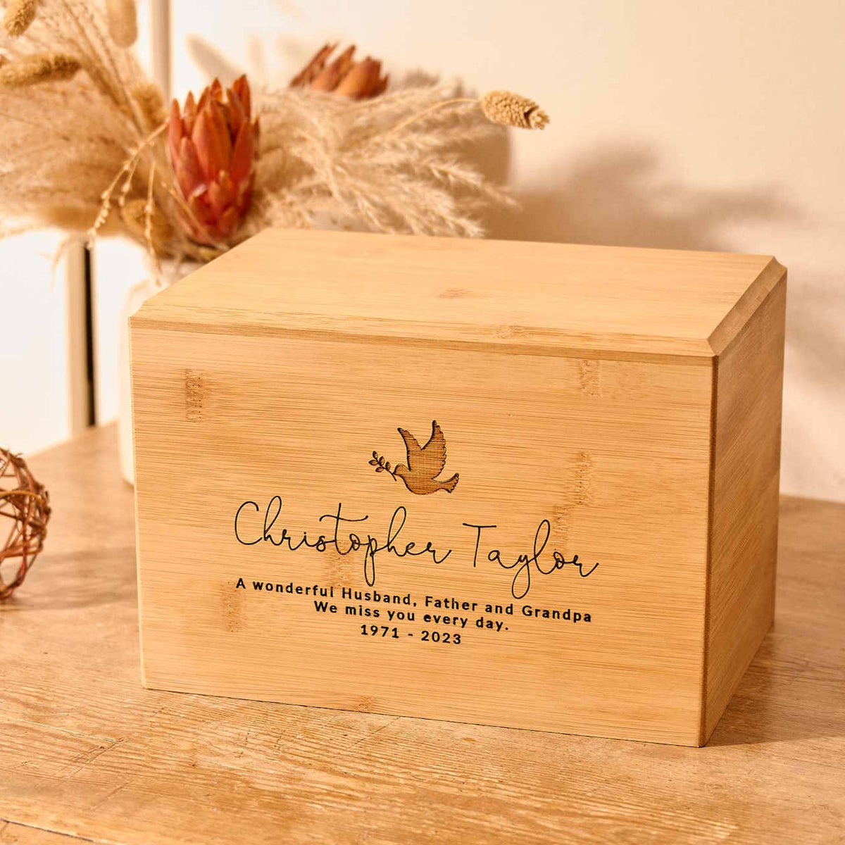 A wooden cremation urn. The urn is rectangular and made of light wood. It has an engraving showing the outline of a dove in flight, carrying an olive branch in its beak. A personalised engraving underneath the dove reads "Christopher Taylor - A wonderful husband, father and grandpa. We miss you every day - 1971-2023"