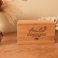 A video showing A personalised cremation urn for a child sits on a wooden table. The urn is rectangular and is engraved on the front face with the words "Amelia - If love could have saved you, you would have lived forever"