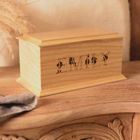 Video shows a wooden cremation urn for ashes sitting on a wooden table. The urn is rectangular and made of light wood, with bevelled edges around the lid and bottom face. It is engraved with the name Emily in capital letters. The letters have a floral design.