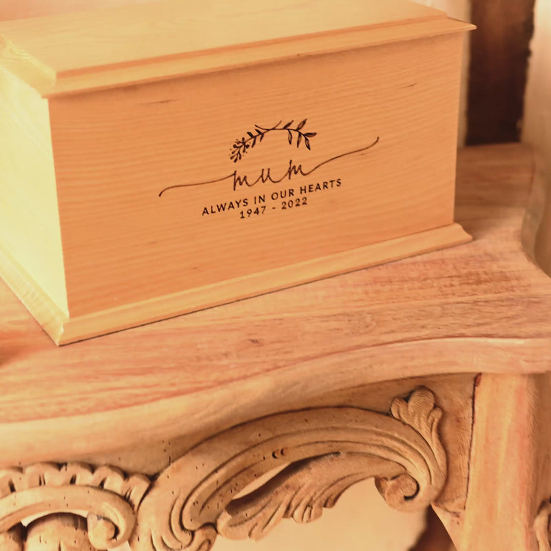 A video showing a wooden cremation urn for ashes from different angles. The urn is made of light wood. It has bevelled edges around the lid and bottom face. It is engraved a simple design showing two crossed laurel leaves. Under the leaves the engraving reads: "Mum - Always in our hearts 1947 - 2022"