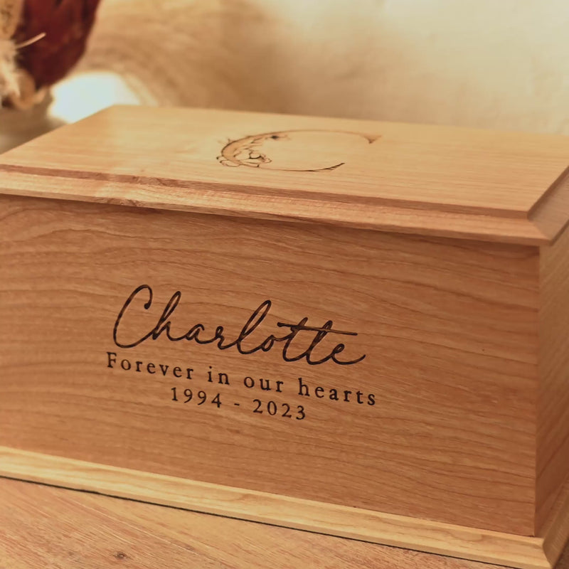 A wooden cremation urn sits on a wooden table. It is a personalised urn. The urn is rectangular and is engraved on the front face with the words "Charlotte - Forever in our hearts 1994 - 2023". The top face of the urn is engraved with a capital letter C with a floral design.