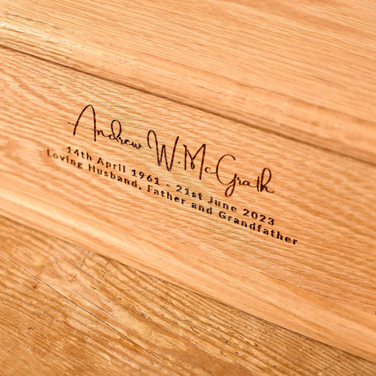 A close up of an engraving on a personalised cremation urn. The urn is made of a light wood. The engraving reads "Andrew W. McGrath - 14th April 1961 - 21st June 2023 - Loving Husband, Father and Grandfather"