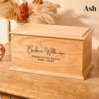 A wooden cremation urn. The urn is rectangular and is made of ash and has a personalised engraving which reads "Barbara Williamson - Always in our hearts - 1945-2023"