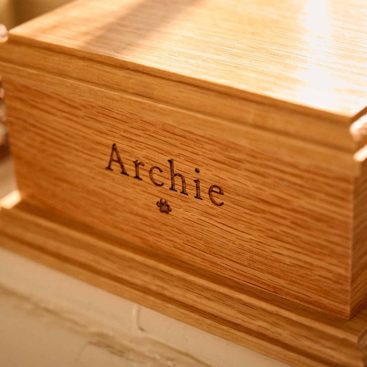 A pet urn. The cremation urn is wooden and sitting on a windowsill.  It has the name Archie engraved on the front face and a paw print. It is a dog urn or a cat urn.