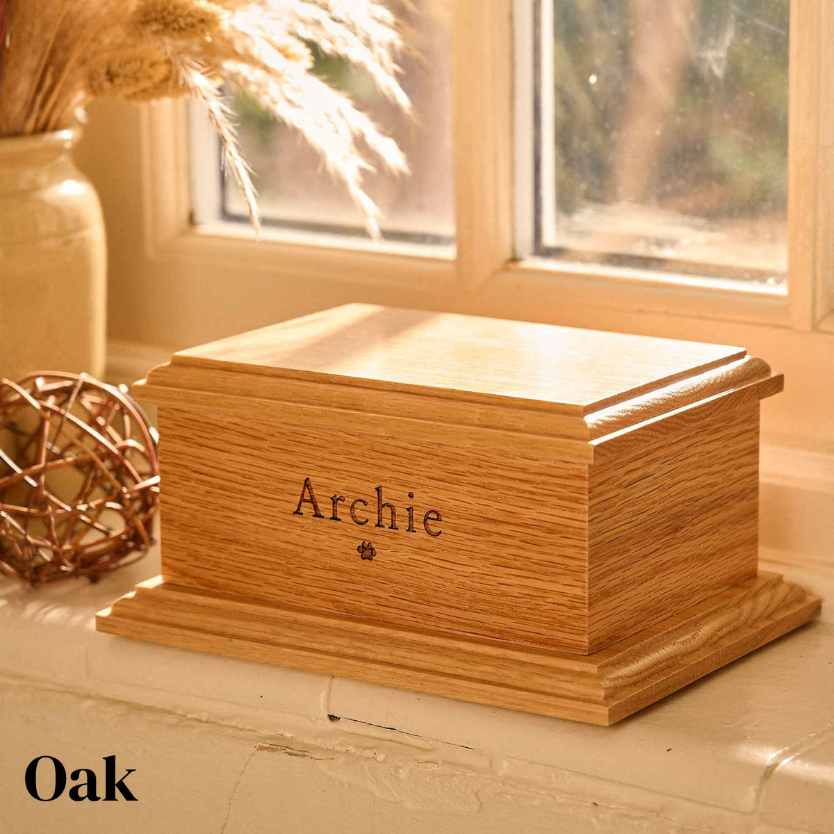 An oak pet cremation urn. The oak urn sits on a windowsill. It has the name Archie and a pawprint engraved on it.