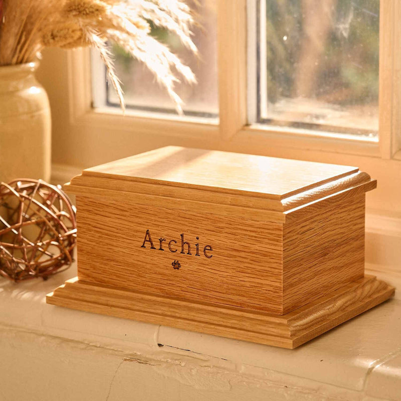 A pet urn. The cremation urn is wooden and sitting on a windowsill.  It has the name Archie engraved on the front face and a paw print. It is a dog urn or a cat urn.