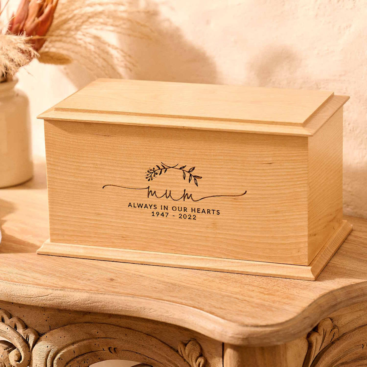 A wooden cremation urn for ashes sits on a wooden table. The urn is rectangular and made of light wood, with bevelled edges around the lid and bottom face. It is engraved a simple design showing two crossed laurel leaves. Under the leaves the engraving reads: "Mum - Always in our hearts 1947 - 2022"