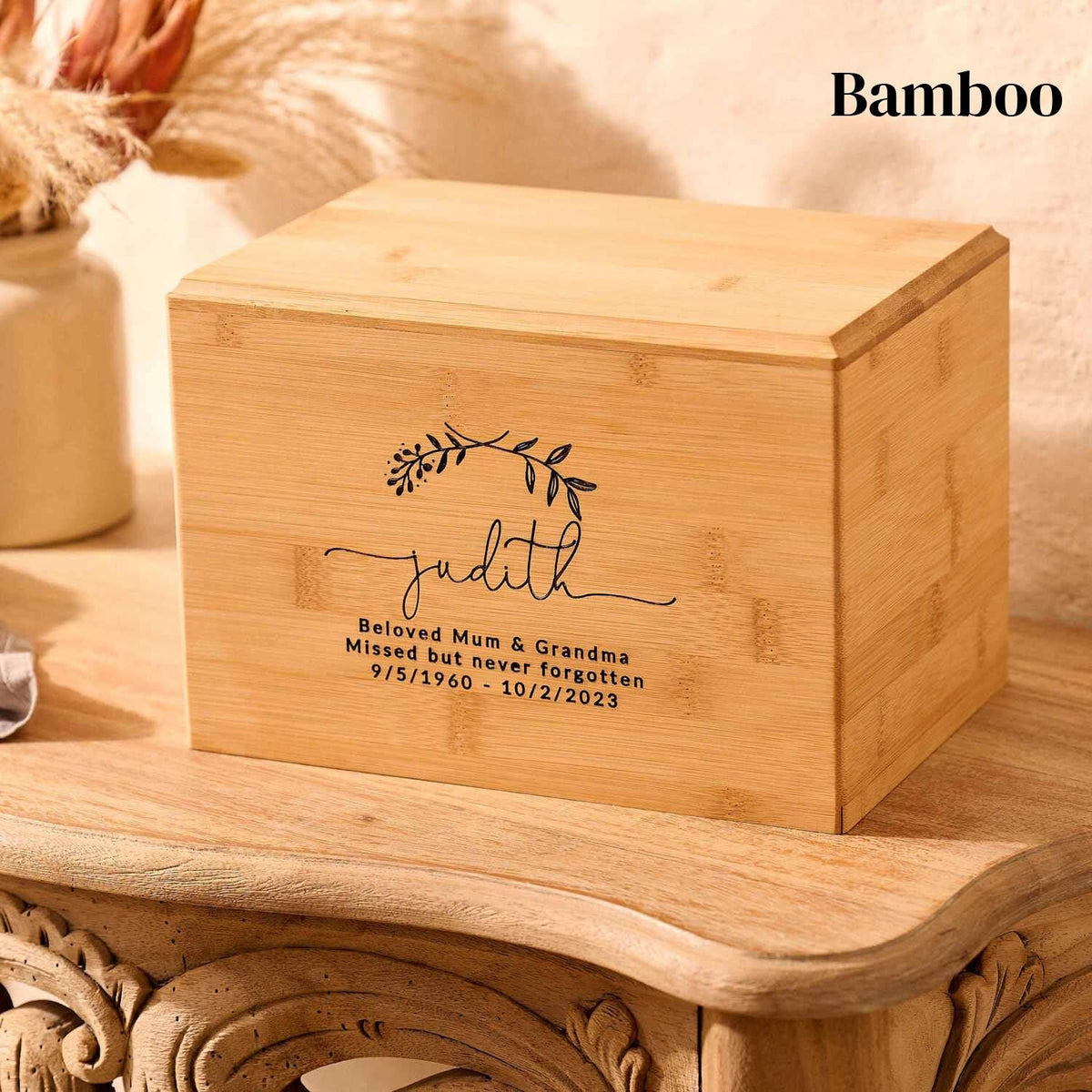 A wooden cremation urn. The urn is rectangular and is made of bamboo. It has a personalised engraving which shows two crossed laurel leaves and a message: "Judith, Beloved Mum & Grandma. Missed but never forgotten. 9/5/1960 - 10/2/23