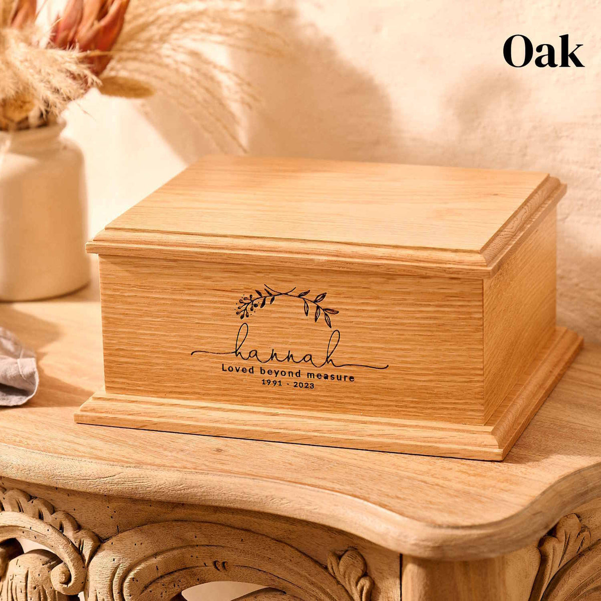 A wooden cremation urn for ashes. The urn is rectangular and is made of oak. Underneath an engraving of two crossed laurel leaves is the message: hannah, loved beyond measure 1991 -2023.