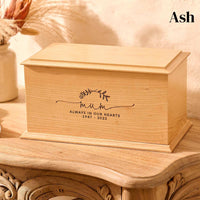 A wooden cremation urn for ashes. The cremation urn is rectangular and is made of ash. It is engraved with a simple design showing two crossed laurel leaves. Under the leaves the engraving reads: "Mum - Always in our hearts 1947 - 2022"