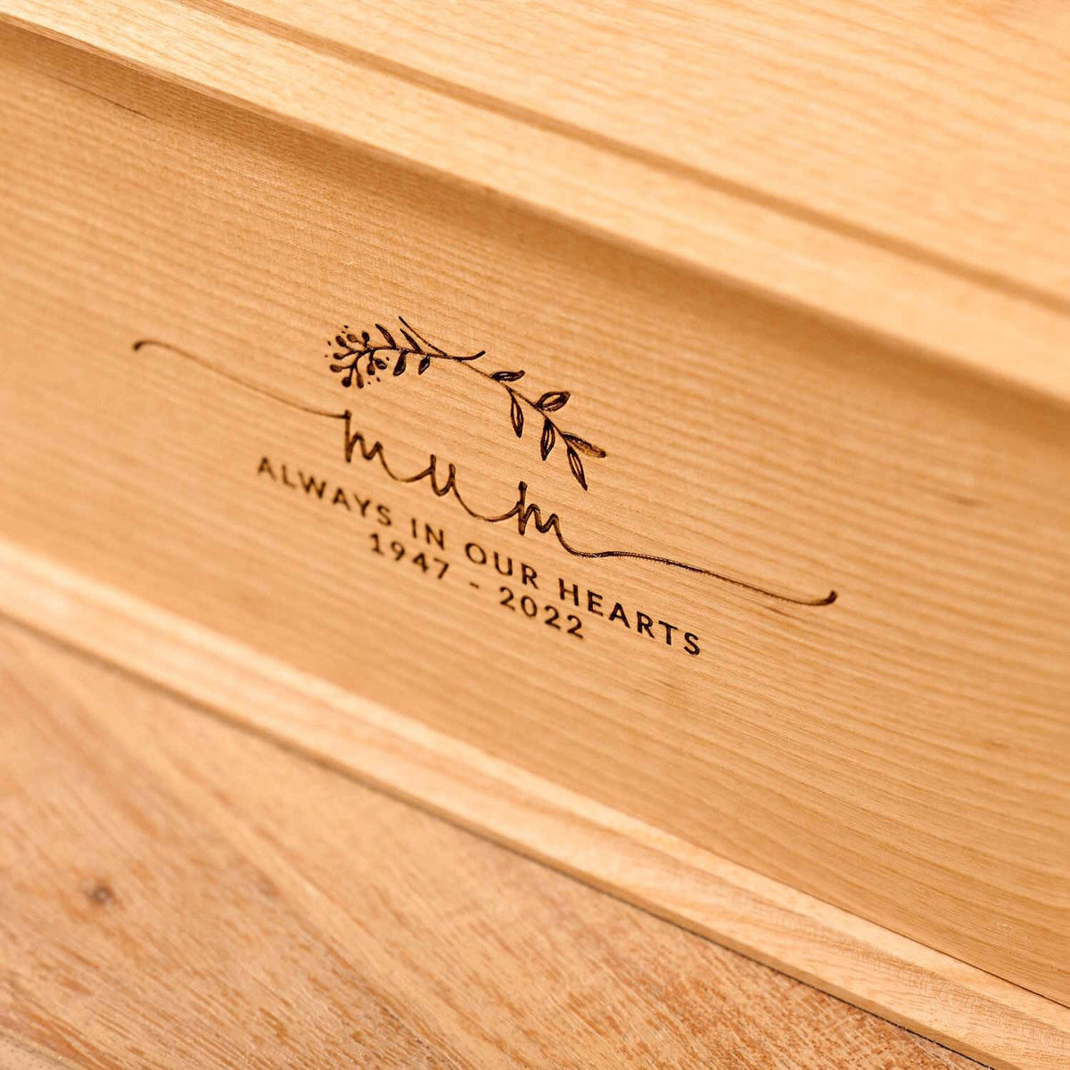 A close up of a wooden cremation urn for ashes. The urn is rectangular and made of light wood, with bevelled edges around the lid and bottom face. It is engraved a simple design showing two crossed laurel leaves. Under the leaves the engraving reads: "Mum - Always in our hearts 1947 - 2022"