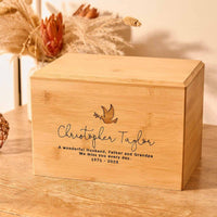 A wooden cremation urn. The urn is rectangular and made of light wood. It has an engraving showing the outline of a dove in flight, carrying an olive branch in its beak. A personalised engraving underneath the dove reads "Christopher Taylor - A wonderful husband, father and grandpa. We miss you every day - 1971-2023"