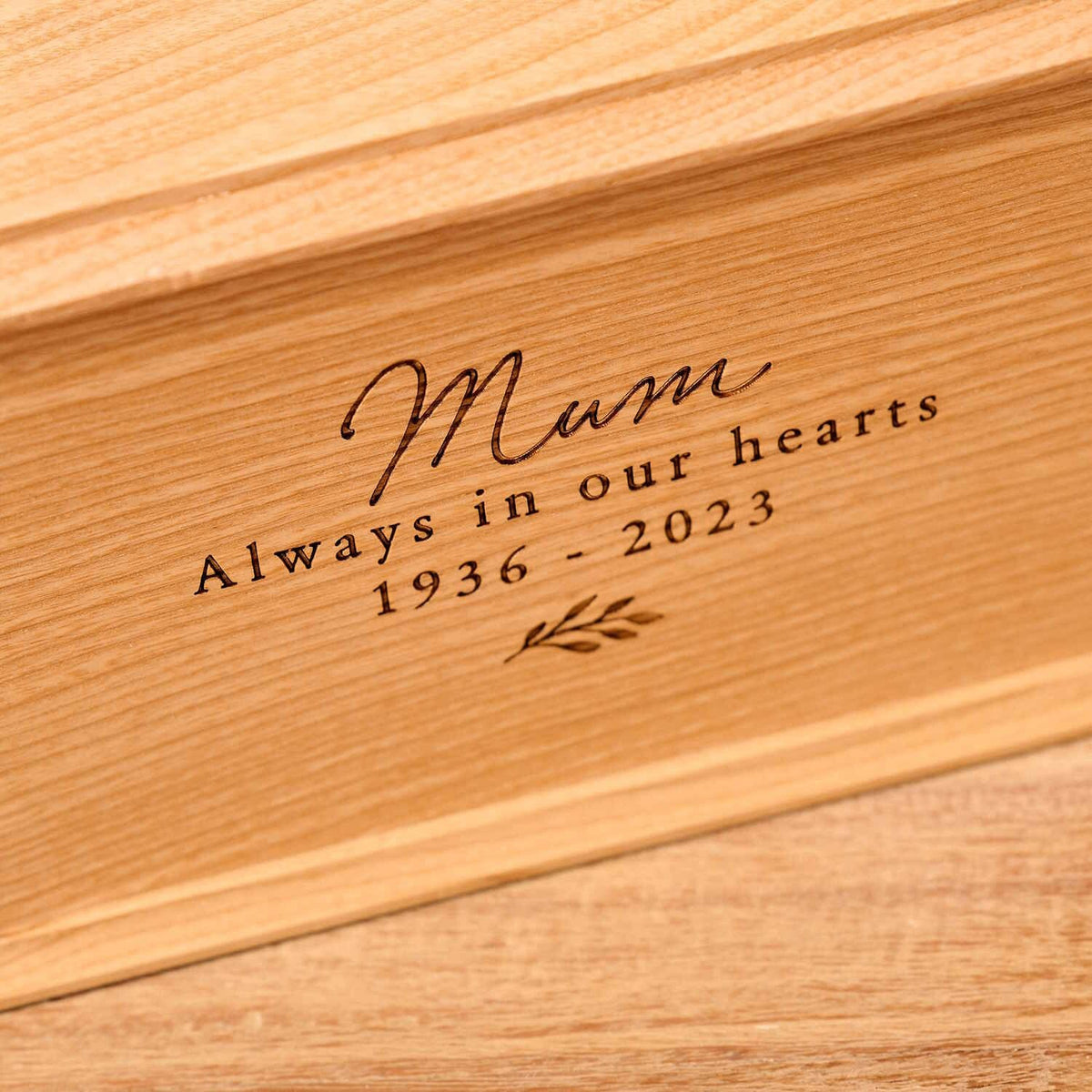 Close up of cremation urn, with bevelled edges around the lid and bottom face. It is engraved on the front face with the words "mum - always in our heards - 1936-2023" and a small leaf motif underneath the next. Urn for mum's ashes.