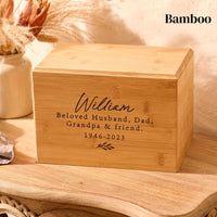 A bamboo cremation urn sitting on a wooden table. The urn is engraved with the words" William, beloved husband, dad, grandpa and friend - 1946-2023" and has a small leaf motif underneath the text