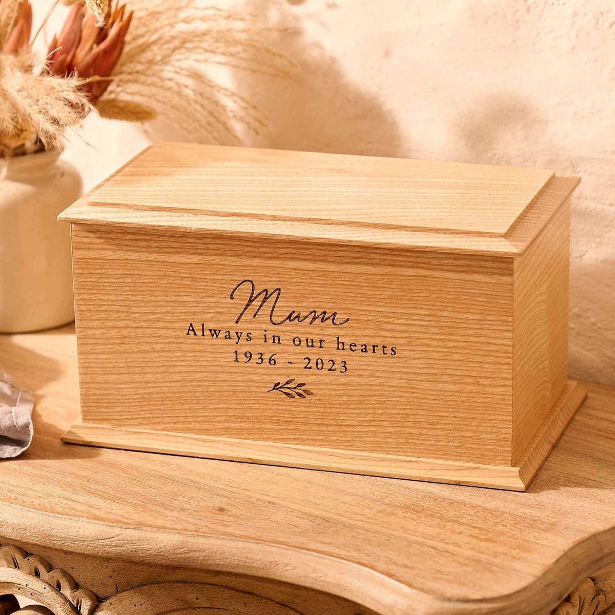 A wooden cremation urn sits on a wooden table. The urn is rectangular and made of ash wood, with bevelled edges around the lid and bottom face. It is engraved on the front face with the words "mum - always in our heards - 1936-2023" and a small leaf motif underneath the next. Urn for mum's ashes.