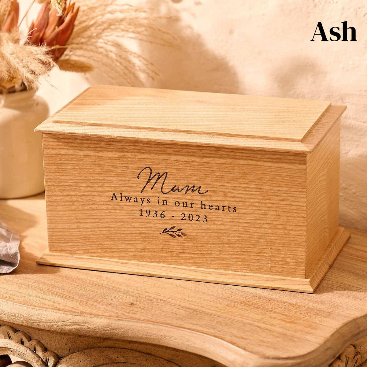 A wooden cremation urn sits on a wooden table. The urn is rectangular and made of ash wood, with bevelled edges around the lid and bottom face. It is engraved on the front face with the words "mum - always in our heards - 1936-2023" and a small leaf motif underneath the next. Engraved urn for mum's ashes.