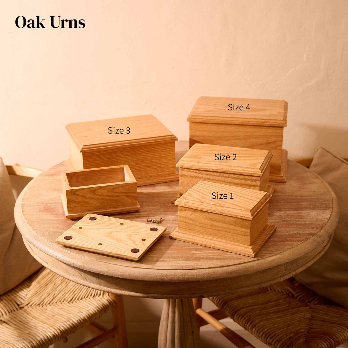 Four oak cremation urns of different sizes on a round wooden table. The urns are all rectangular and are all made of oak wood. they have bevelled edges on the top and bottom faces. There are two identical small urns, which are labelled "Size 1". One of these small urns has its lid removed, showing the inside of the urn (which is empty). The lid and four screws sit on the table. The medium urn is labelled "Size 2". The large urn is lablled "Size 3". Both the medium and large urns are shown with their lids