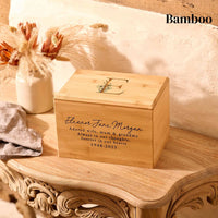 A wooden cremation urn sits on a wooden table. It is a personalised urn made of bamboo. The urn is rectangular and is engraved on the front face with a name and dates of birth and death. The top face of the urn is engraved with a capital letter E with a floral design.