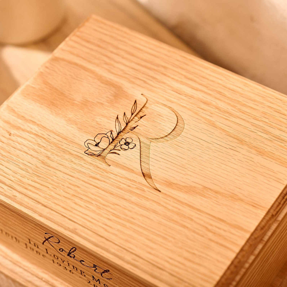 A close up of a personalised cremation urn. The photograph shows the detail of an engraved letter R, with a floral design. It is a wooden urn.