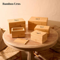 Five wooden cremation urns of different sizes on a round wooden table. The urns are all rectangular and are all made of bamboo wood. they have simple bevelled edges on the top face. There are two identical small urns, which are labelled "Size 1". One of these small urns has its lid removed, showing the inside of the urn (which is empty). The lid and and one screw sits on the table. The medium urn is labelled "Size 2". The large urn is labelled "Size 3". The extra large urn is labelled "Size 4".