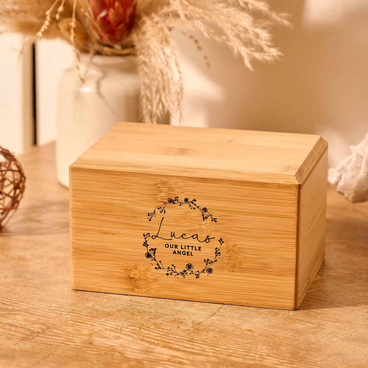 An personalised cremation urn for a child sits on a wooden table. The urn is rectangular and is engraved on the front face with the words "Lucas - Our Little Angel". It is an urn for a stillborn baby, an urn for a child or an urn for a miscarriage. The text is encased with a floral wreath.