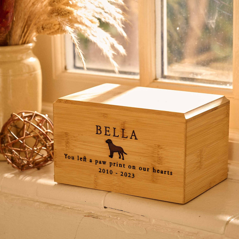 A pet urn for dog ashes. The cremation urn is wooden and sitting on a windowsill.  It has the name Bella engraved on it and has a picture of a dog. It is a Labrador urn