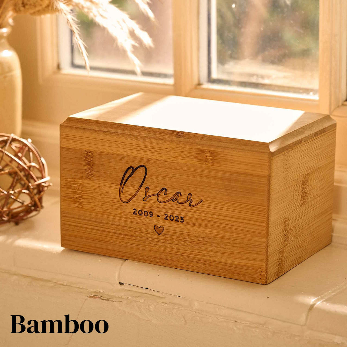 A bamboo cat urn or dog urn. It says "oscar" on it. It is sitting on a windowsill. Personalised pet urn.