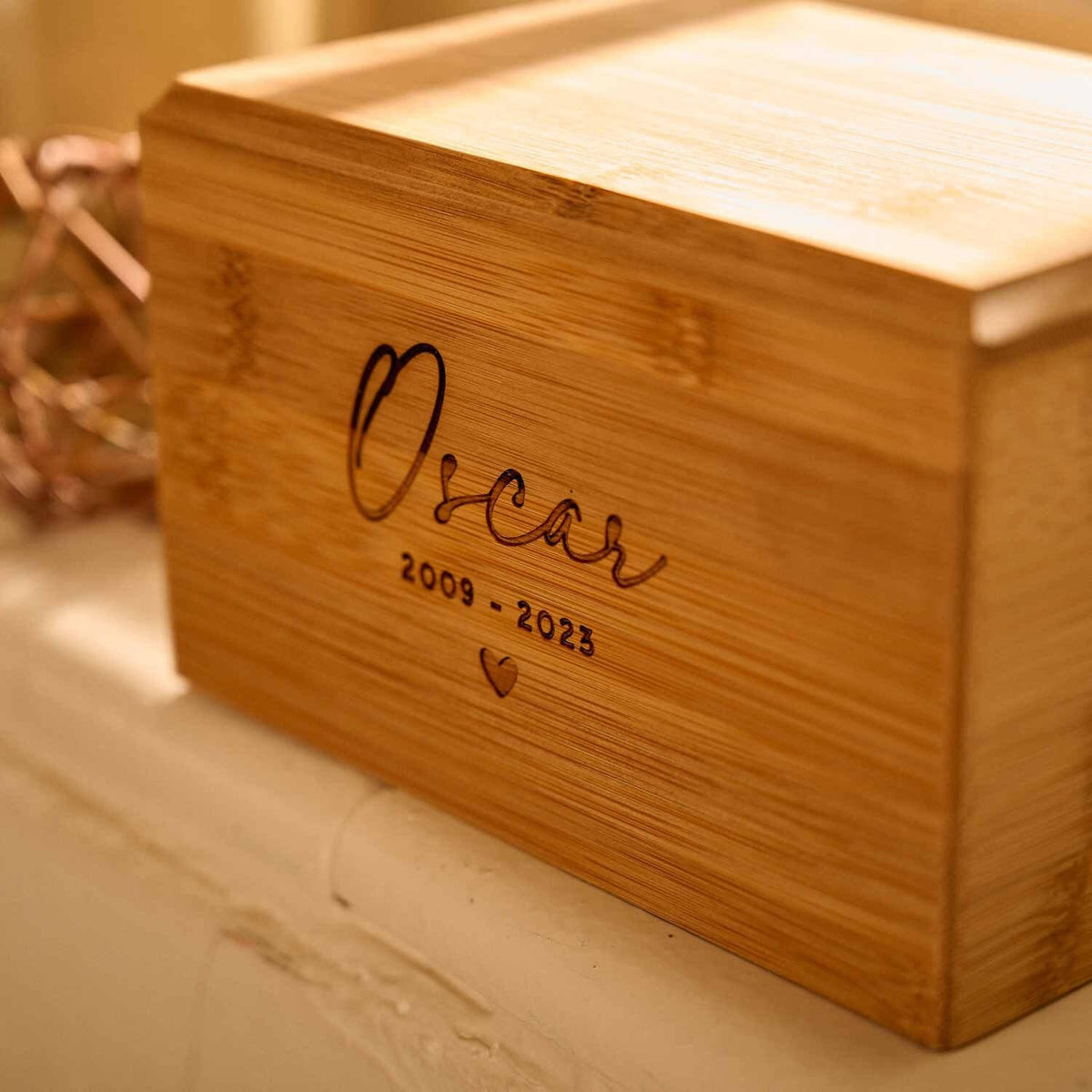 A cremation urn for pet ashes sitting on a windowsill. The urn is engraved with "Oscar 2009 - 2023" and a heart.