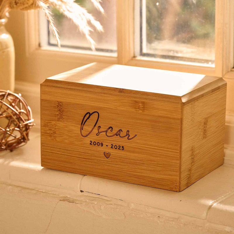 A cremation urn for pet ashes sitting on a windowsill. The urn is engraved with "Oscar 2009 - 2023" and a heart.