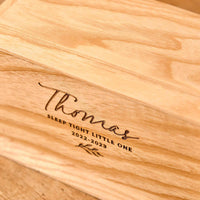 A personalised cremation urn for a child sits on a wooden table. The urn is rectangular and is engraved on the front face with the words "Thomas - Sleep Tight Little One 2022-2023". The cremation urn has a leaf motif.