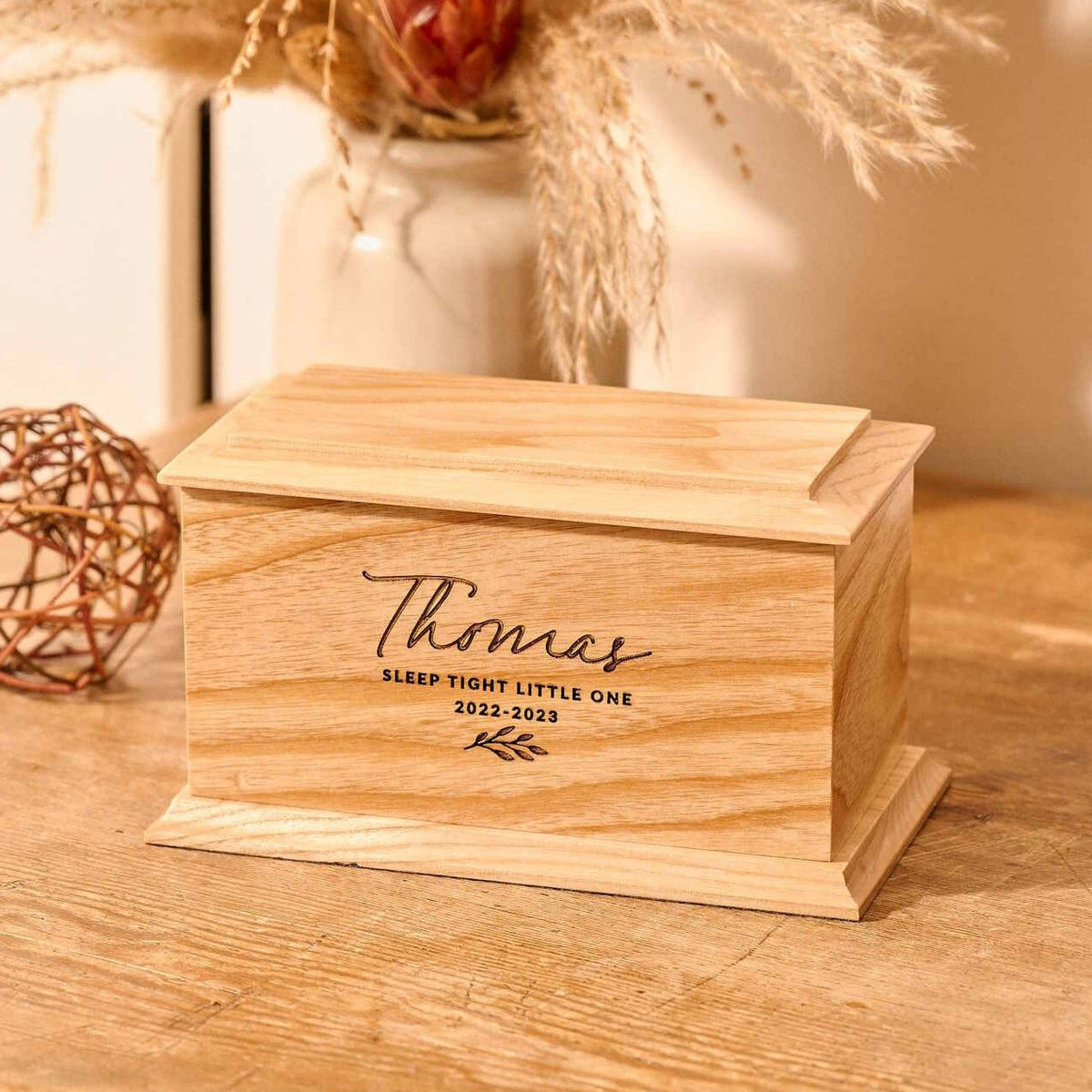 A personalised cremation urn for a child sits on a wooden table. The urn is rectangular and is engraved on the front face with the words "Thomas - Sleep Tight Little One 2022-2023". The cremation urn has a leaf motif.