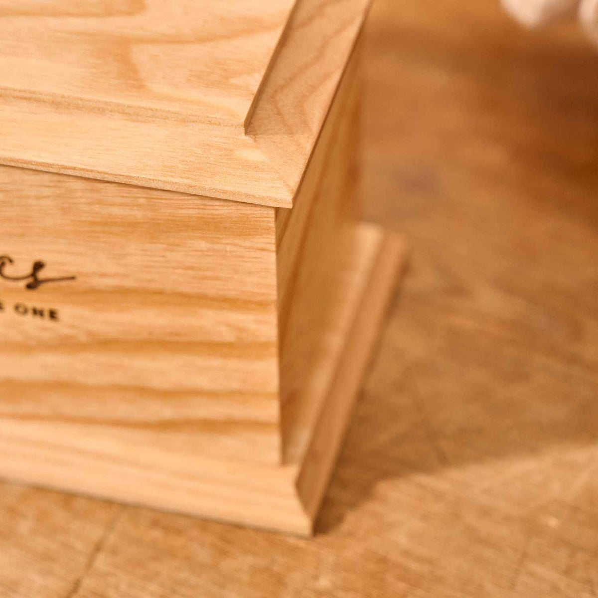 A close up of detail on a cremation urn. The urn is rectangluar and is made of a light wood. The photograph is focused on bevelling detail around the top face 