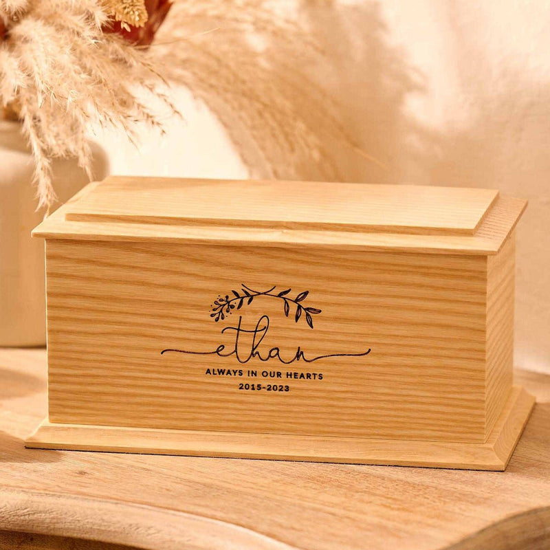 A personalised cremation urn for a child sits on a wooden table. The urn is rectangular and is engraved on the front face with the name Ethan. It is an urn for a stillborn baby, an urn for a child or an urn for a miscarriage.