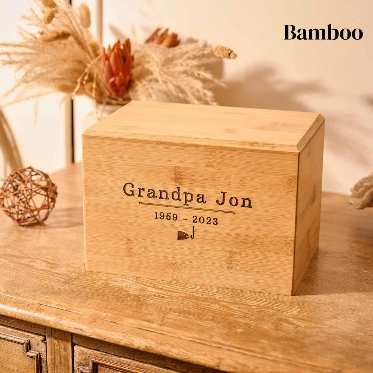 A wooden cremation urn for ashes. The cremation urn is rectangular and is made of bamboo. It is engraved with the words "Grandpa Jon - 1959 -2023" and a small picture of a fish and a fishing hook