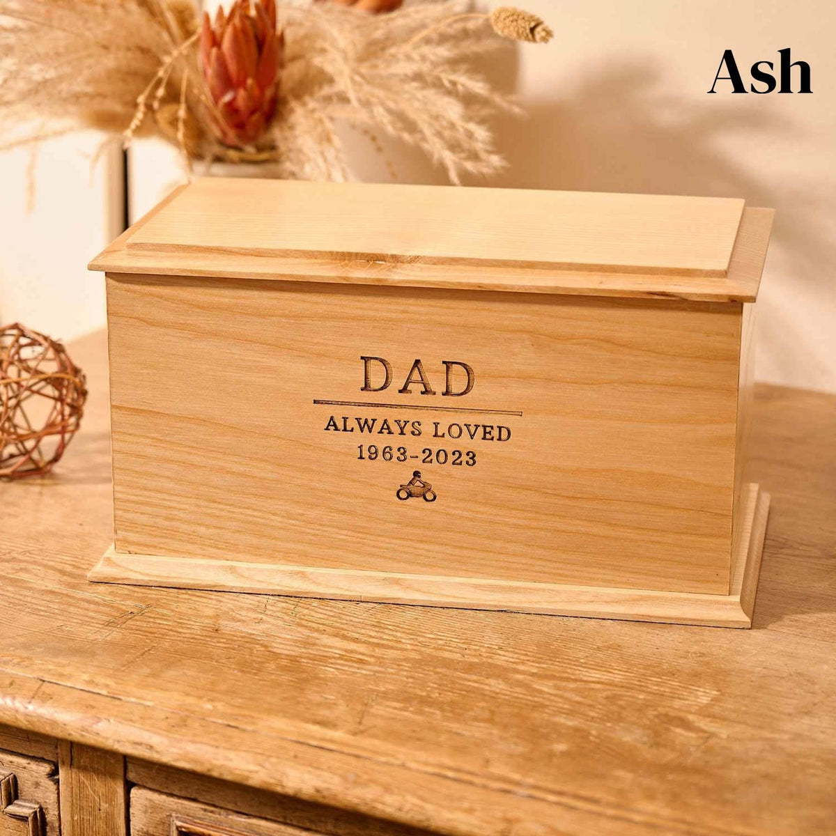 A wooden cremation urn for ashes. The cremation urn is rectangular and is made of ash. It is engraved with the words "Dad - Always Loved 1963 - 2023" and a small picture of a person sitting on a motorbike. 