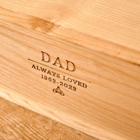 A close up of a wooden cremation urn for ashes. The urn is engraved with the words "Dad - Always Loved 1963 - 2023" and a small picture of a person sitting on a motorbike.