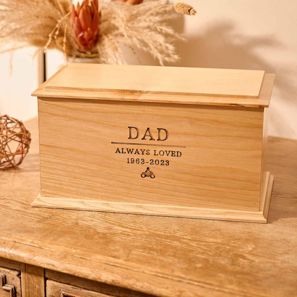 A wooden cremation urn for ashes sits on a wooden table. The urn is rectangular and made of light wood, with bevelled edges around the lid and bottom face. It is engraved with the words "Dad - Always Loved 1963 - 2023" and a small picture of a person sitting on a motorbike.