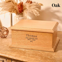 A wooden cremation urn for ashes. The cremation urn is rectangular and is made of oak. It is engraved with the words "Thomas - Our darling son - 2004-2023" and a small picture of a games console controller 