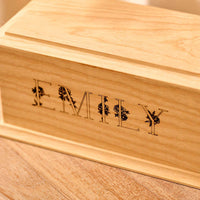 A wooden urn for cremation ashes. The urn is rectangular and made of light wood, with bevelled edges around the lid and bottom face. It is engraved with the name Emily in capital letters. The letters have a floral design.