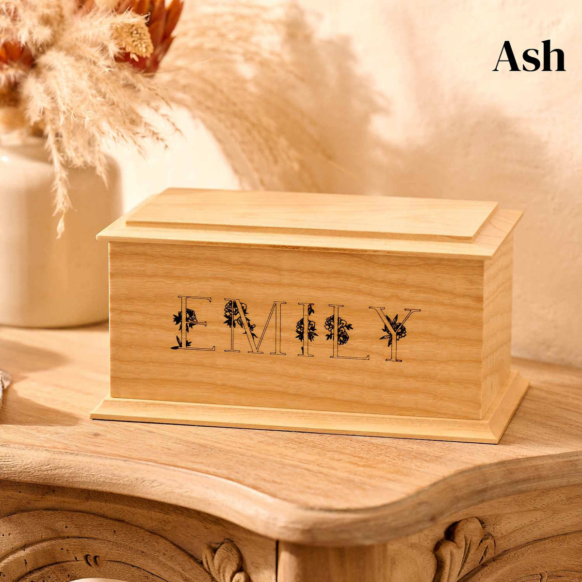 A wooden cremation urn for ashes sits on a wooden table. The urn is rectangular and made of ash wood, with bevelled edges around the lid and bottom face. It is engraved with the name Emily in capital letters. The letters have a floral design.