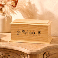 A wooden cremation urn for ashes sits on a wooden table. The urn is rectangular and made of light wood, with bevelled edges around the lid and bottom face. It is engraved with the name Emily in capital letters. The letters have a floral design.