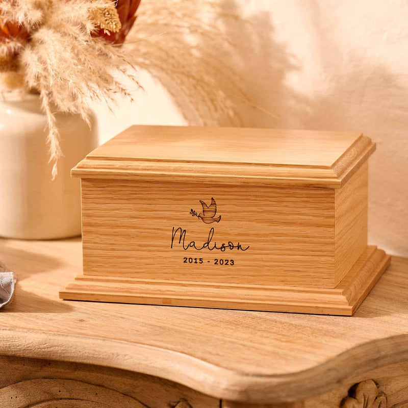 A personalised wooden cremation urn for a child sits on a wooden table. The urn is rectangular and is engraved on the front face with the name Madison. It is an urn for a stillborn baby, an urn for a child or an urn for a miscarriage.