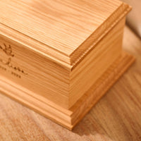 A personalised wooden cremation urn for a child sits on a wooden table. The urn is rectangular and is engraved on the front face with the name Madison. It is an urn for a stillborn baby, an urn for a child or an urn for a miscarriage. 