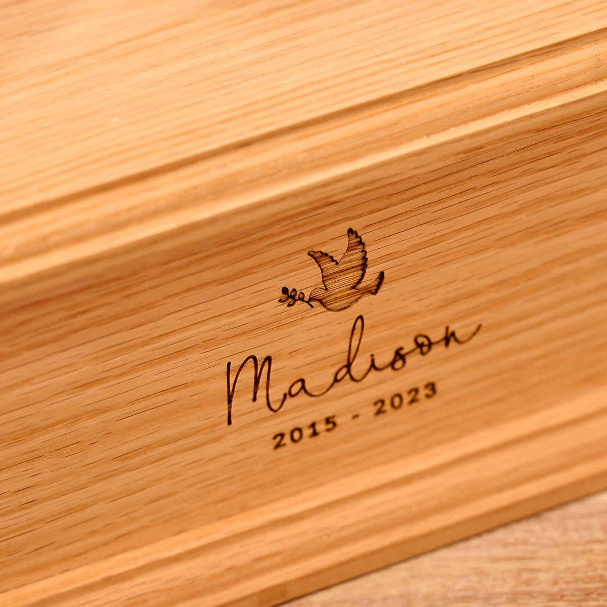 A personalised wooden cremation urn for a child sits on a wooden table. The urn is rectangular and is engraved on the front face with the name Madison. It is an urn for a stillborn baby, an urn for a child or an urn for a miscarriage.