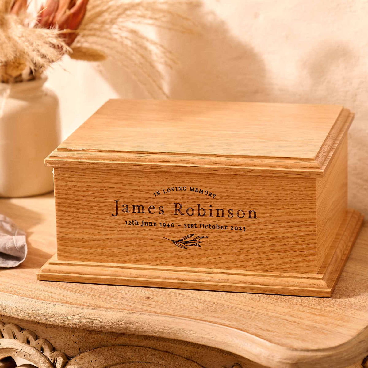 An engraved wooden cremation urn sits on a wooden table. It is an urn for a man. The man's name is James Robinson - this is engraved on the cremation urn.